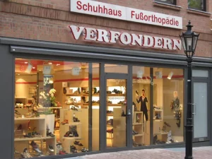De afbeelding toont de voorgevel van Schuhhaus Verfondern, een schoen- en voetorthopediewinkel met grote glazen etalages vol schoenen en een rood-wit uithangbord boven de ingang.