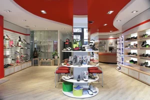 De afbeelding toont het midden van een moderne schoenwinkel, met een rond presentatiemeubel vol schoenen en tijdschriften, omringd door wandrekken met meer schoenen en een rood-wit plafondontwerp.