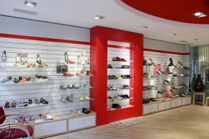 De afbeelding toont het interieur van een moderne schoenwinkel met witte lamellenwanden en glazen planken, waarop tal van dames- en herenschoenen zijn uitgestald. In het midden zorgt een rood nis-element voor een opvallend accent, terwijl onder de wanden witte ladekasten staan op een lichte houten vloer.