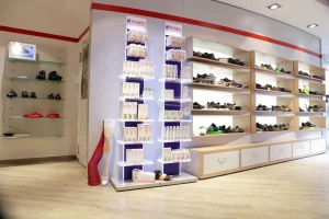 De afbeelding toont het interieur van een moderne schoenwinkel: rechts lange, verlichte houten schappen vol schoenen, en in het midden een display met GEHWOL-voetverzorgingsproducten.